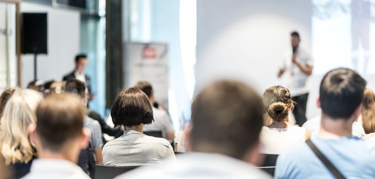 Business and entrepreneurship symposium. Speaker giving a talk at business meeting. Audience in conference hall. Rear view of unrecognized participant in audience.