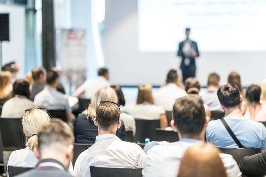 Business and entrepreneurship symposium. Speaker giving a talk at business meeting. Audience in conference hall. Rear view of unrecognized participant in audience.