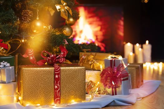 Stack of packed gift boxes under Christmas tree against burning fireplace. Lots of Christmas gifts under the tree. Candles on wooden floor. Focus on golden box!
