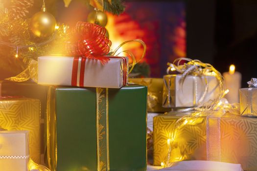 Stack of packed gift boxes under Christmas tree against burning fireplace. Lots of Christmas gifts under the tree. Candles on wooden floor. Focus on gray box!