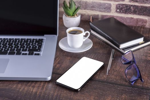 Laptop notebook with blank screen and cup of coffee and notepad pen and books and smart-phone on wooden table. 
Mock up on wooden table with laptop with blank screen, cup of coffee, pen, books and flower.
Focus on smart-phone and glasses.