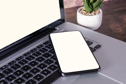 Smartphone white blank screen on laptop keyboard on wooden table with supplies.
