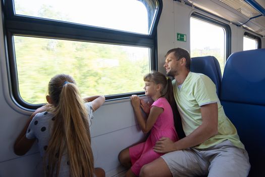 Dad and two daughters in an electric train car, look out the window with enthusiasm