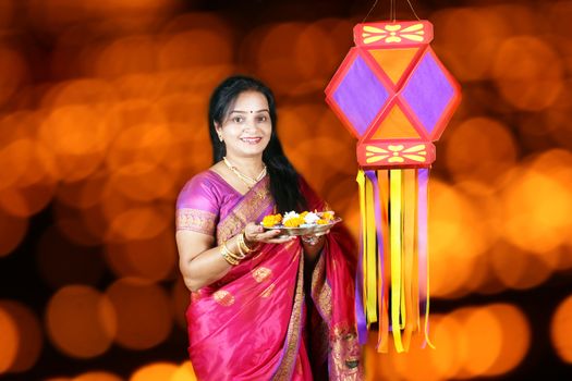 An middle aged happy Indian woman wearing a traditional saree holding a plate of ritual items standing near a sky lantern in Diwali