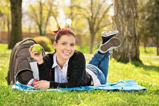 Healthy life with nature around us. Beautiful smiling young girl lying on the grass in the park holding an apple, taking a break from studying.
