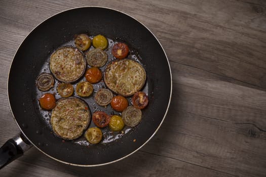 Frying pan with fried aubergine cherry tomatos and onion on a wooden table, top view