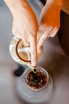 Hot green tea poured out of the teapot into the ice chocolate glass on table.