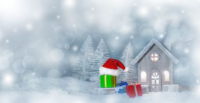 Christmas card with cozy small decorative house , gifts and Santa Claus cap on sleigh on snow over fairy tale winter fir forest background and falling snow . New Year design postcard . Copy space for text