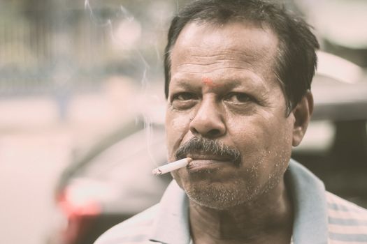 Portrait of a A mid adult senior mature man of Asian and Indian Ethnicity is smoking cigarette. Front View. Close up. Photography taken on 31 May 2019 on World No Tobacco or anti Tobacco Day, in a city street of India, South Asia.