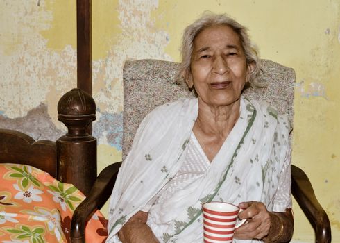 Portrait of smiling Active senior Asian and Indian Ethnicity woman of 82 years sitting on chair holding a cup of coffee resting at home while looking at camera.