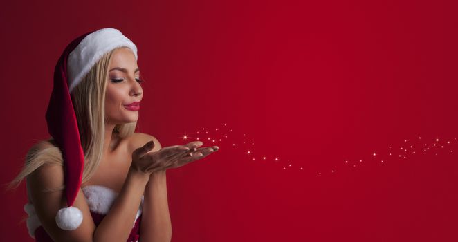 Beautiful sexy woman in santa hat and red dress blowing fairy dust off her palms over red background with copy space