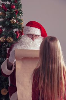 Santa Claus with with list or nice or naugthy list paper with small girl near christmas tree