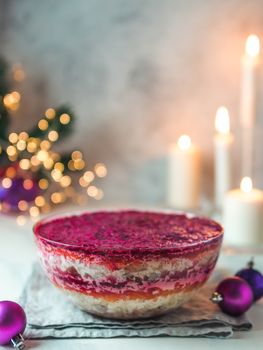 Layered salad herring under a fur coat on festive table, with candle, fir tree and light garland. Traditional russian salad with herring and vegetables in glass bowl. Copy space for text. Vertical.