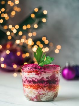 Layered salad herring under a fur coat on festive table, with fir tree and light garland. Traditional russian salad with herring and vegetables in glass jar. Copy space for text. Vertical.