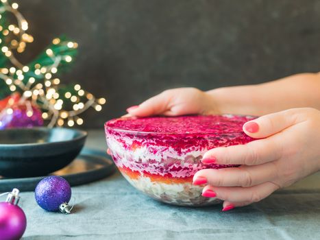 Woman hands with layered salad herring under a fur coat on festive table. Traditional russian salad with herring and vegetables in glass bowl. Copy space for text.