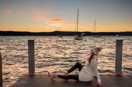 Woman sitting in relaxed pose on the jetty watching the winter sunset.  She is wearing a knitted beanie and sweater dress