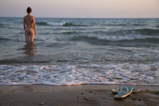Beautiful young woman in bikini on the beach stands in water looking on the sea.