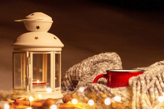 Lantern with a burning candle, spices and a red mug with hot coffee on a snowy wooden table.