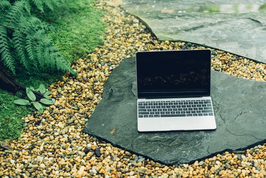 Laptop computer mockup has empty screen outside on stone in garden of coffee shop.