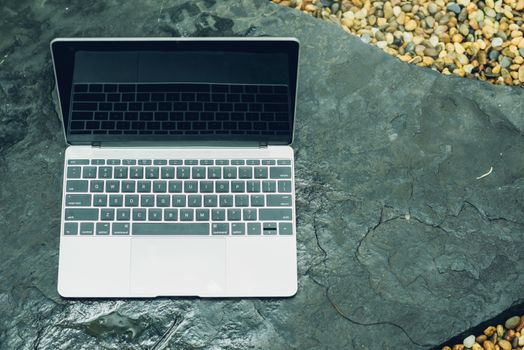 Laptop computer mockup has empty screen outside on stone in garden of coffee shop.