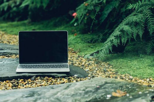 Laptop computer mockup has empty screen outside on stone in garden of coffee shop.