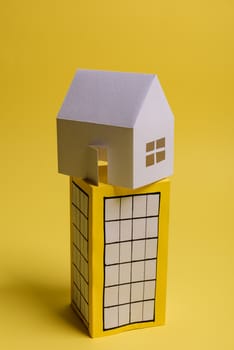 White family paper house over block of flats on yellow background paper. Minimalistic and simple concept, style. Copy space. Vertical orientation.