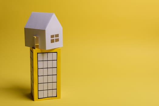 White family paper house over block of flats on yellow background paper. Minimalistic and simple concept, style. Copy space. Horizontal orientation.