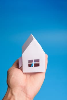 White family paper house in man hand on blue background paper. Minimalistic style. Copy space. View from above. Vertical orientation