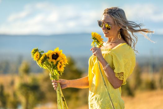 Happy woman holding bright yellow sunflowers, hair blowing in the wind in a rural landscape scene