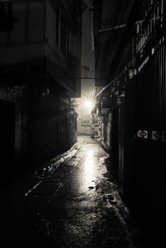 A dark, shadowy and dangerous looking urban back-alley at night time in suburbs Hanoi, Vietnam. Low light reflected on wet pavement from post lamp at the end of long road corner