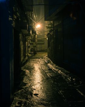 Vintage tone a dark, shadowy and dangerous looking urban back-alley at night time in suburbs Hanoi, Vietnam. Low light reflected on wet pavement from post lamp at the end of long road corner