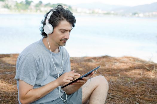 young man holding a tablet with headphones, at the beach