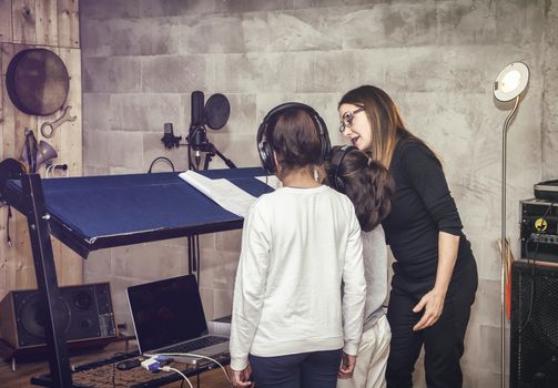 Rear view of two unrecognizable children in headphones with female acting or singing teacher in recording studio