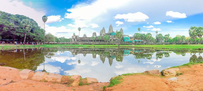 Panorama lakeside Angkor Wat facade under construction and palm tree reflection under summer cloud blue sky with unidentified tourists. A temple complex in Cambodia, world largest religious monument
