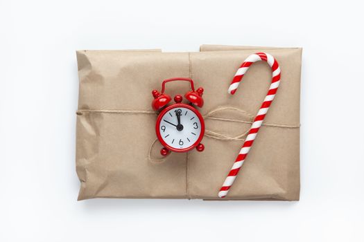 Christmas gift wrapped in brown craft paper, tied with scourge, decorated with cane candy and small analog clock on white background. Minimal style. Top view. Do it yourself, celebration, eco concept.