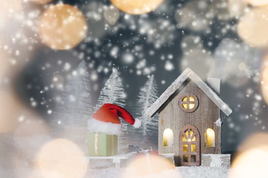 Christmas card with cozy small decorative house , gifts and Santa Claus cap on sleigh on snow over fairy tale winter fir forest background and falling snow . New Year design postcard . Copy space for text
