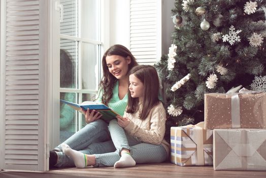 Mother and daughter reading a book sitting on the floor near christmas tree