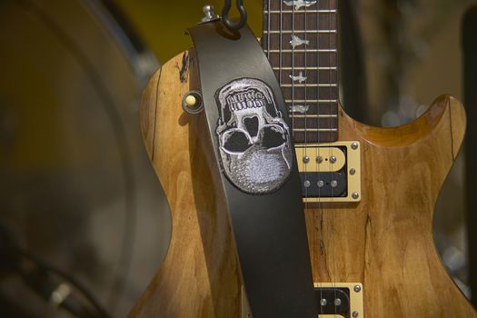 Beautiful detail of an excellent light wood electric guitar, with the shoulder strap placed in front with a skull embroidered. The handle and the strings are clearly visible while the background is blurred to focus the attention on the guitar itself.
