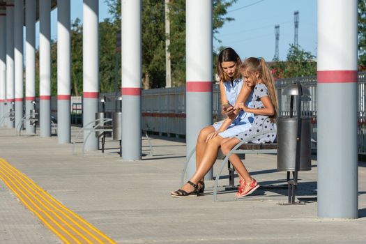 Mom and daughters wait for the train and look at the phone on the empty platform of the railway station