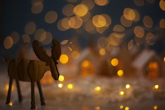 Christmas wooden reindeer over small glowing toy houses village garland over bokeh lights background with copy space for text