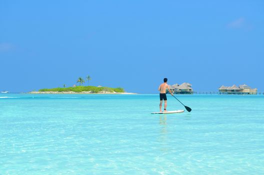 Young asian man paddle board rowing on sunny tropical paradise island with blue  sea water ocean in Maldives island.