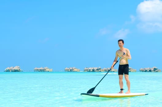 Young asian man paddle board rowing on sunny tropical paradise island with blue  sea water ocean in Maldives island.