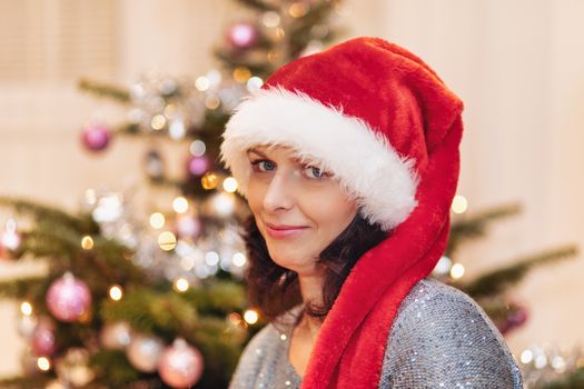 Beautiful middle age woman in red party santa hat sitting near the Christmas tree