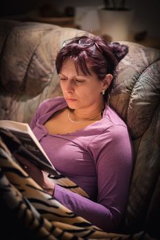 middle-aged woman is reading a book at home with the lamp on. Authentic photo, real people in real situation