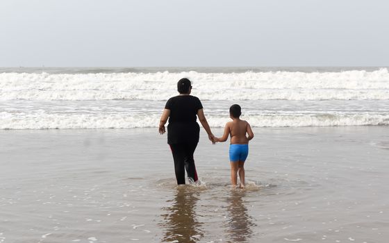 Rear View of Mother and son standing on a tropical beach in evening during sunset. The child admires his mom,s faith. Happy Mother’s day background concept. Goa, India, South Asia Pacific In Summer.