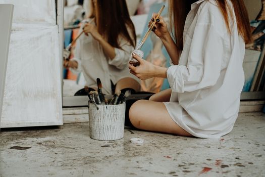 Woman painter sitting on the floor in front of mirror and drawing. Art studio interior. Drawing supplies, oil paints, artist brushes