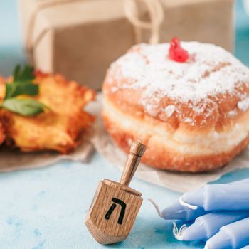 Jewish holiday Hanukkah concept and background. Hanukkah food doughnuts and potatoes pancakes latkes, giftbox, candle and traditional spinnig dreidl on blue background. Copy space for text.