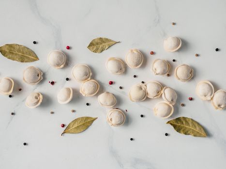 Pattern of frozen uncooked russian pelmeni with peppercorns and bay leaves on white marble table. Creative layout of dumplings. Beautiful scattered raw dumplings. Top view or flat lay.