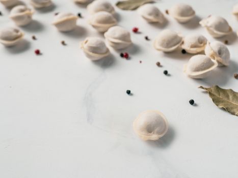Pattern of frozen uncooked russian pelmeni with peppercorns and bay leaves on white marble table. Creative layout of dumplings. Beautiful scattered raw dumplings. Top view or flat lay. Copy space