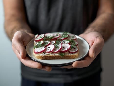 Hands takes plate with vegan sandwich. Healthy appetiezer - whole wheat bread toast with plant-based soft cheese, radish, dill and black sesame